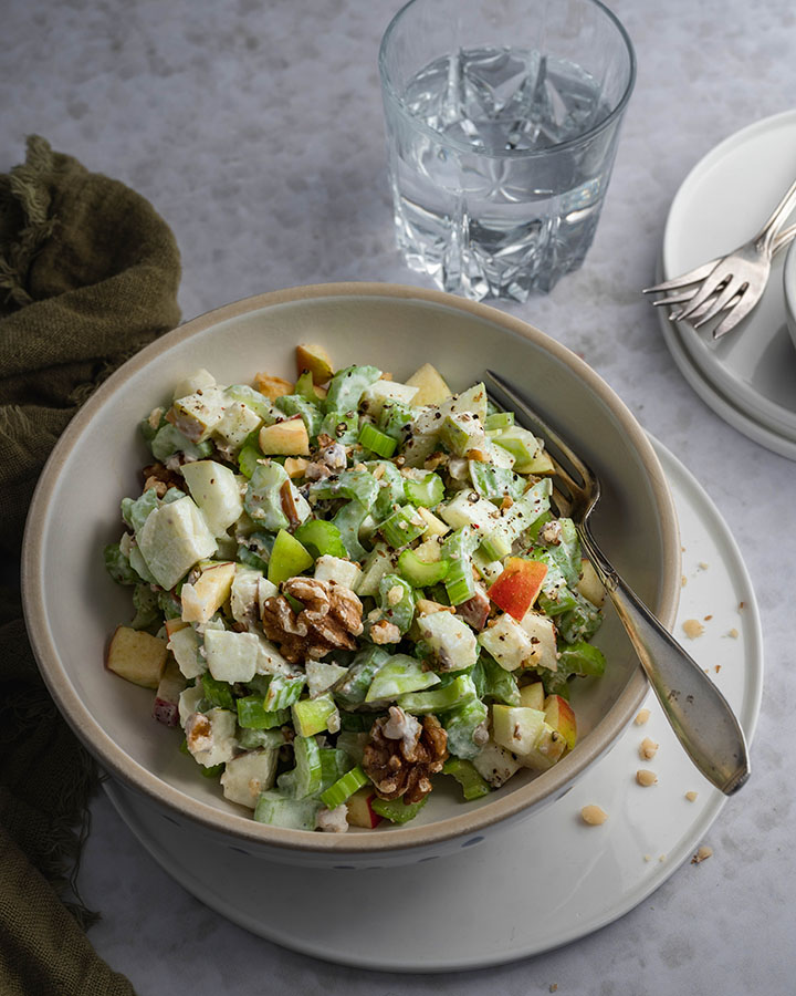 Eine Schüssel mit Waldorfsalat steht auf einem Tisch. Daneben liegt Besteck und ein Glas Wasser.