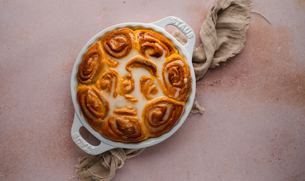 Eine Backform mit Kürbis-Zimtschnecken und Zuckerguss stehen auf einem Tisch.