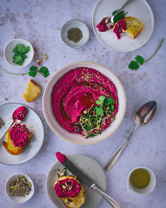 Eine Schüssel mit Rote Bete Humus steht auf einem Tisch. Daneben sind kleine Teller mit Brot zu Dippen.