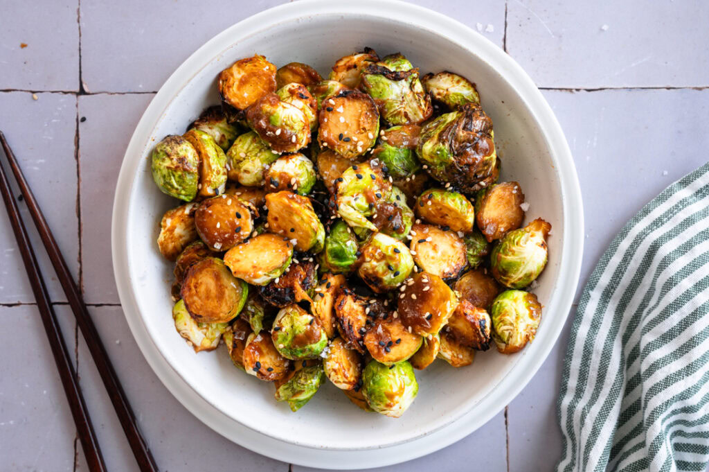 Eine weiße Schüssel mit knusprigem Rosenkohl aus dem Airfryer steht auf einem Fliesentisch.