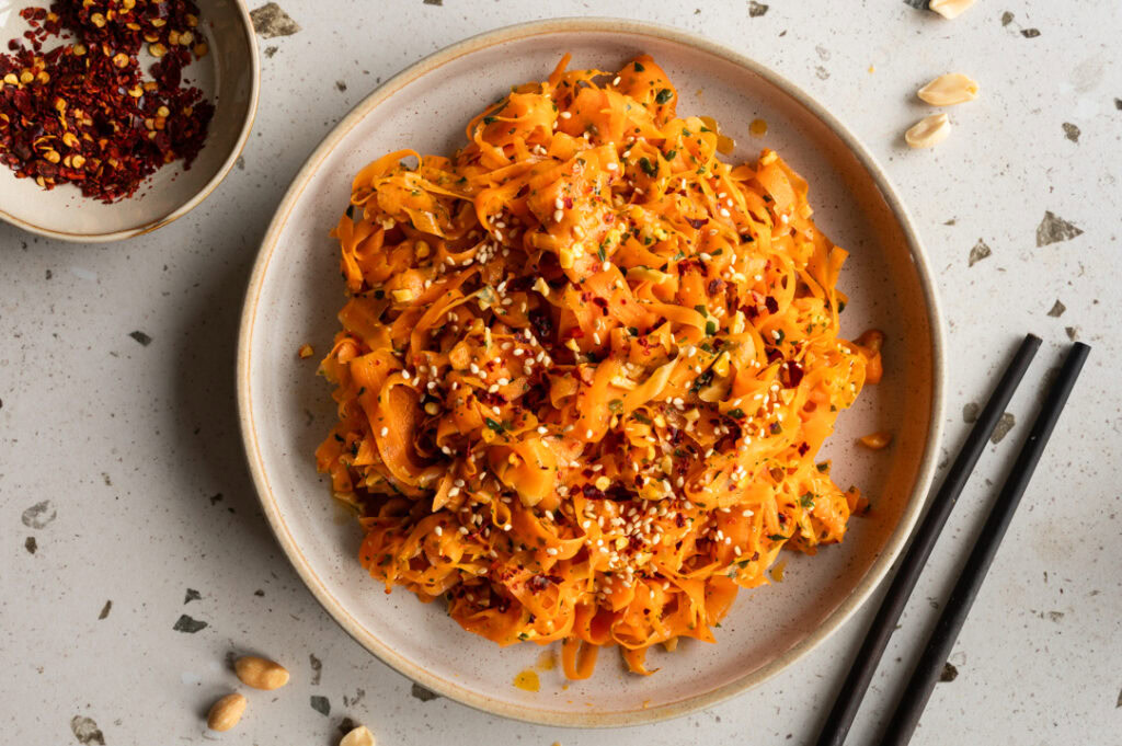 Ein Teller mit asiatischem Karottensalat und eine kleine Schale Chiliflocken stehen auf dem Tisch.