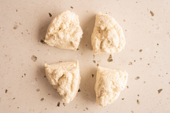 Der Teig ist in 4 Teile aufgeteilt und die Teile liegen auf einem Tisch.