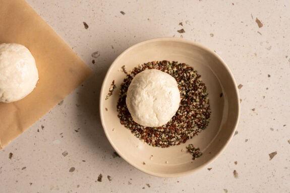 Ein Brötchen wird kopfüber in eine Schüssel getunkt, in der eine Mischung aus Leinsamen, Hanfsamen und Chiasamen ist.