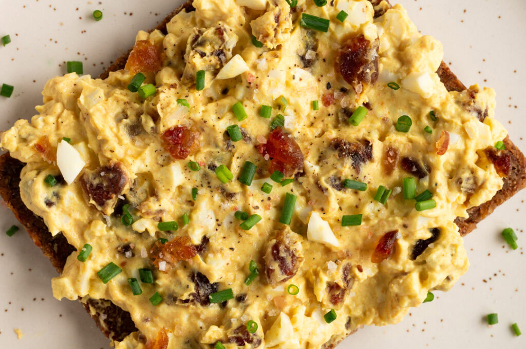 Eine Scheibe Roggenbrot bestrichen mit Eiersalat mit Curry und Datteln liegt auf einem Teller.