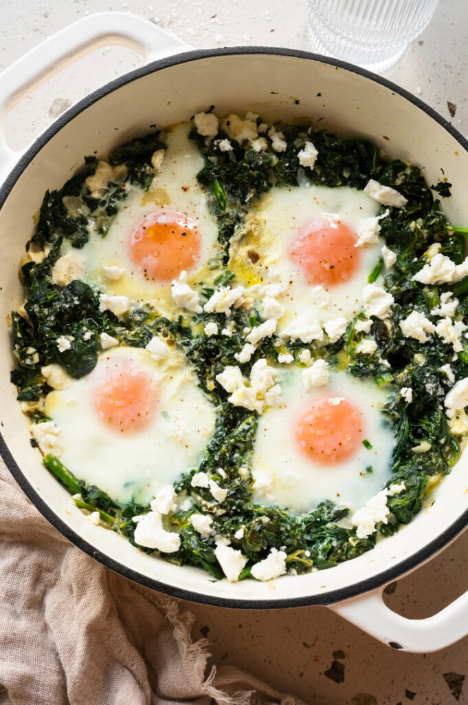 Eine tiefe, weiße Pfanne mit Spinat, Feta und Eiern steht auf einem Tisch.