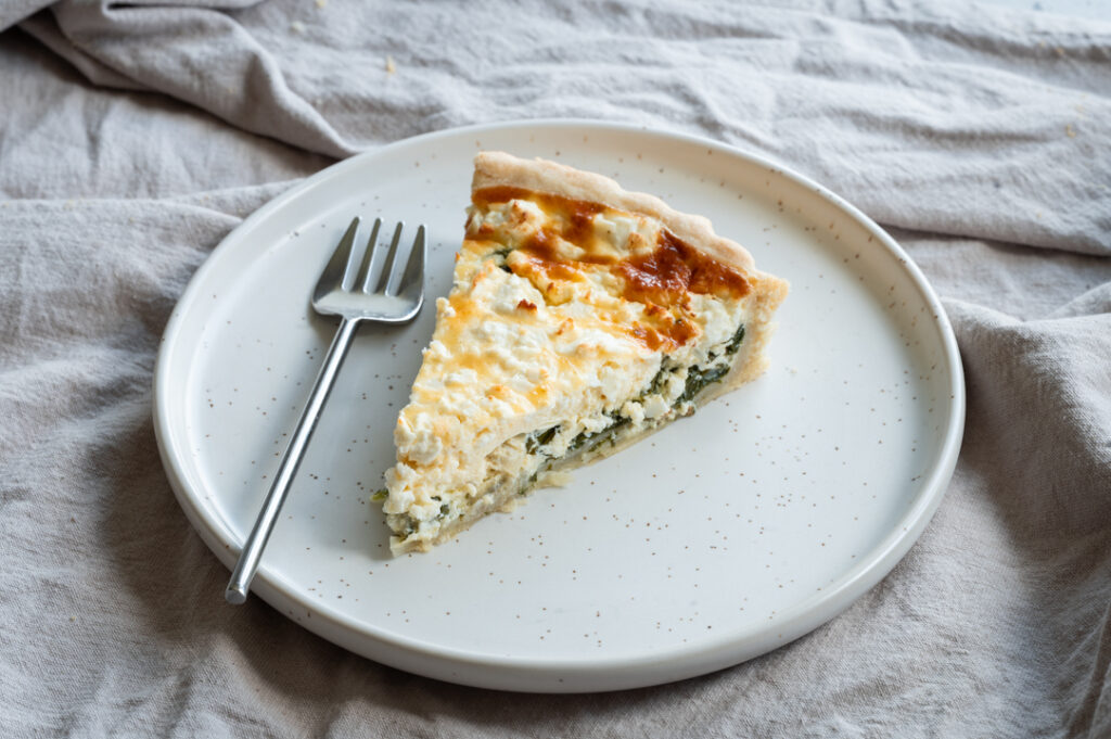 Ein Stück einer Quiche mit Spinat und Feta liegt auf einem Teller.