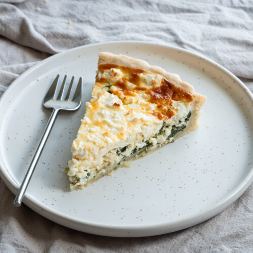 Ein Stück einer Quiche mit Spinat und Feta liegt auf einem Teller.